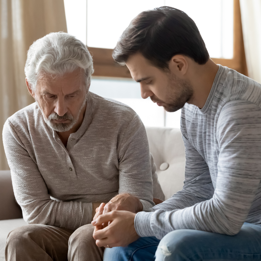 A Quiet Moment of Support - American Hero Properties We Buy Houses Young man comforting an older man by holding his hands as they sit close together on a couch, both looking emotional and reflective.
