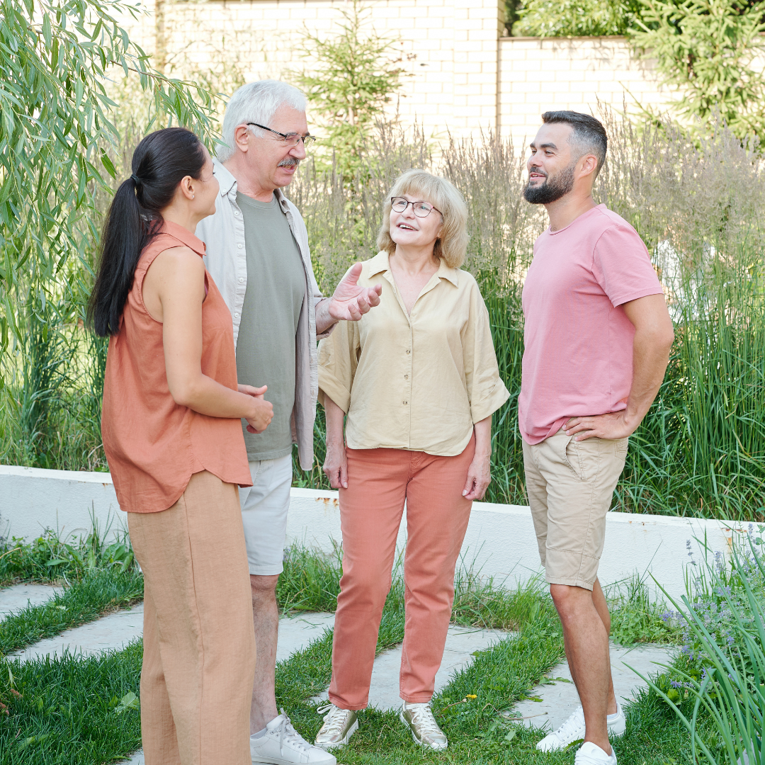 Coming Together for a Family Decision - American Hero Properties Buys Houses for Cash Alt text: Four adults standing outdoors in a landscaped garden, casually chatting and smiling. Two older individuals are speaking with two younger people, suggesting a family conversation.