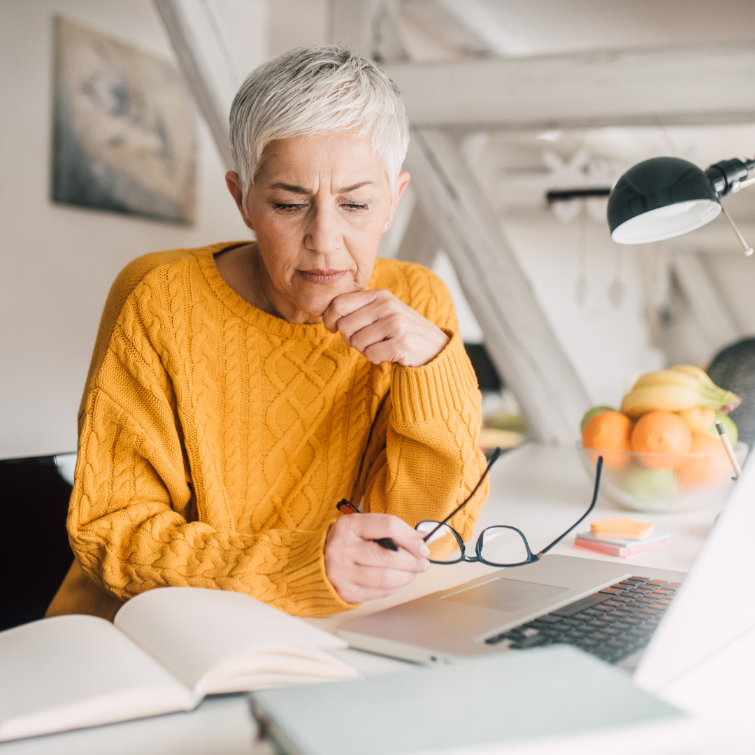 Navigating the Uncertainty of Inherited Property Timelines Older woman in a cozy sweater looking thoughtfully at a laptop, surrounded by paperwork, suggesting she’s researching or managing an inherited property.