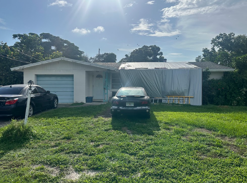 Florida Home Sold As-Is With Roof Damage Single-story Florida home with tarp covering part of the roof, two parked cars, and a patchy grass front yard under bright sunlight.
