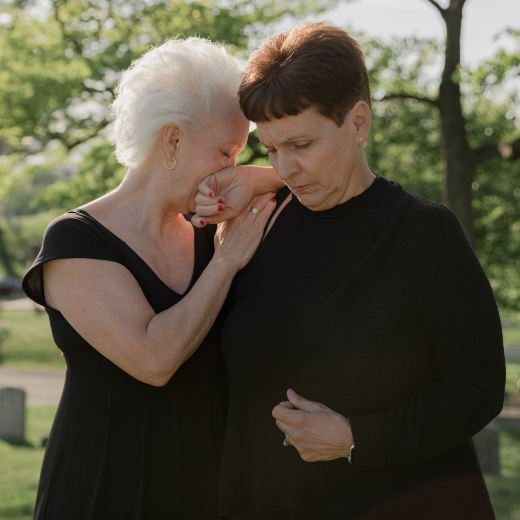 Two older women grieving together outdoors, embracing during an emotional moment in a cemetery